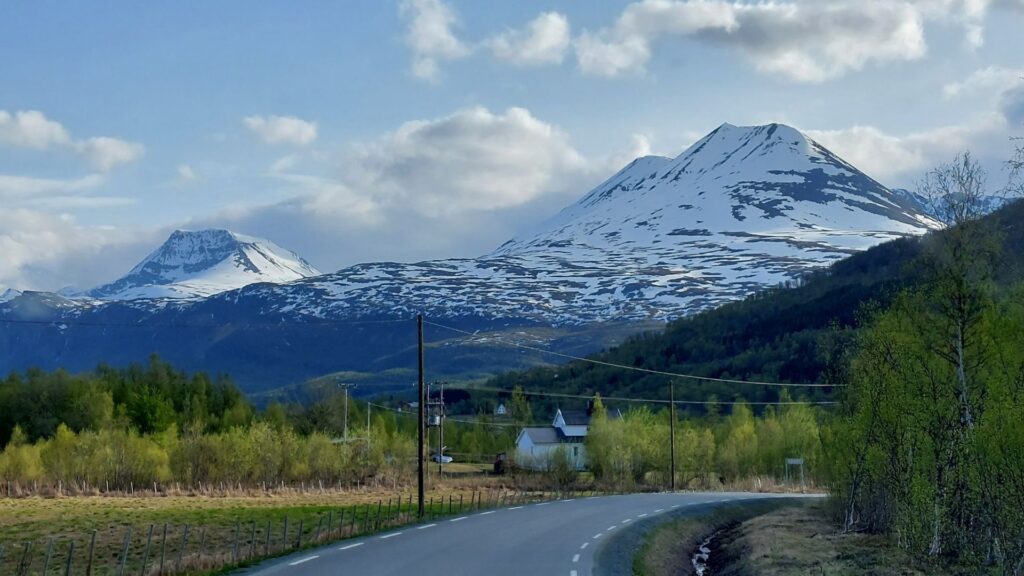 Vista a la carretera y con las montanas al fondo, cubiertas con nieve. Se ve el cielo y las nubes blancas.