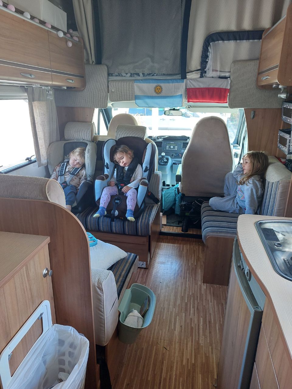 interior de la autocaravana capuchina. dos ninos durmiendo y otra nina sentada en la silla. dos banderas de polonia y argentina colgadas.